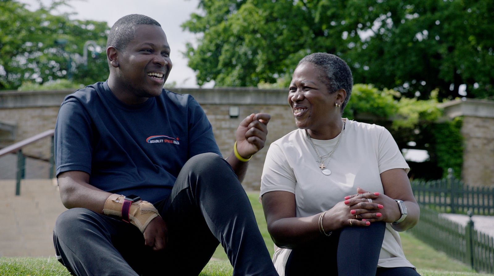 A man wearing a Disability Sports Coach t-shirt and his mum are both sat on a lawn while talking and smiling.
