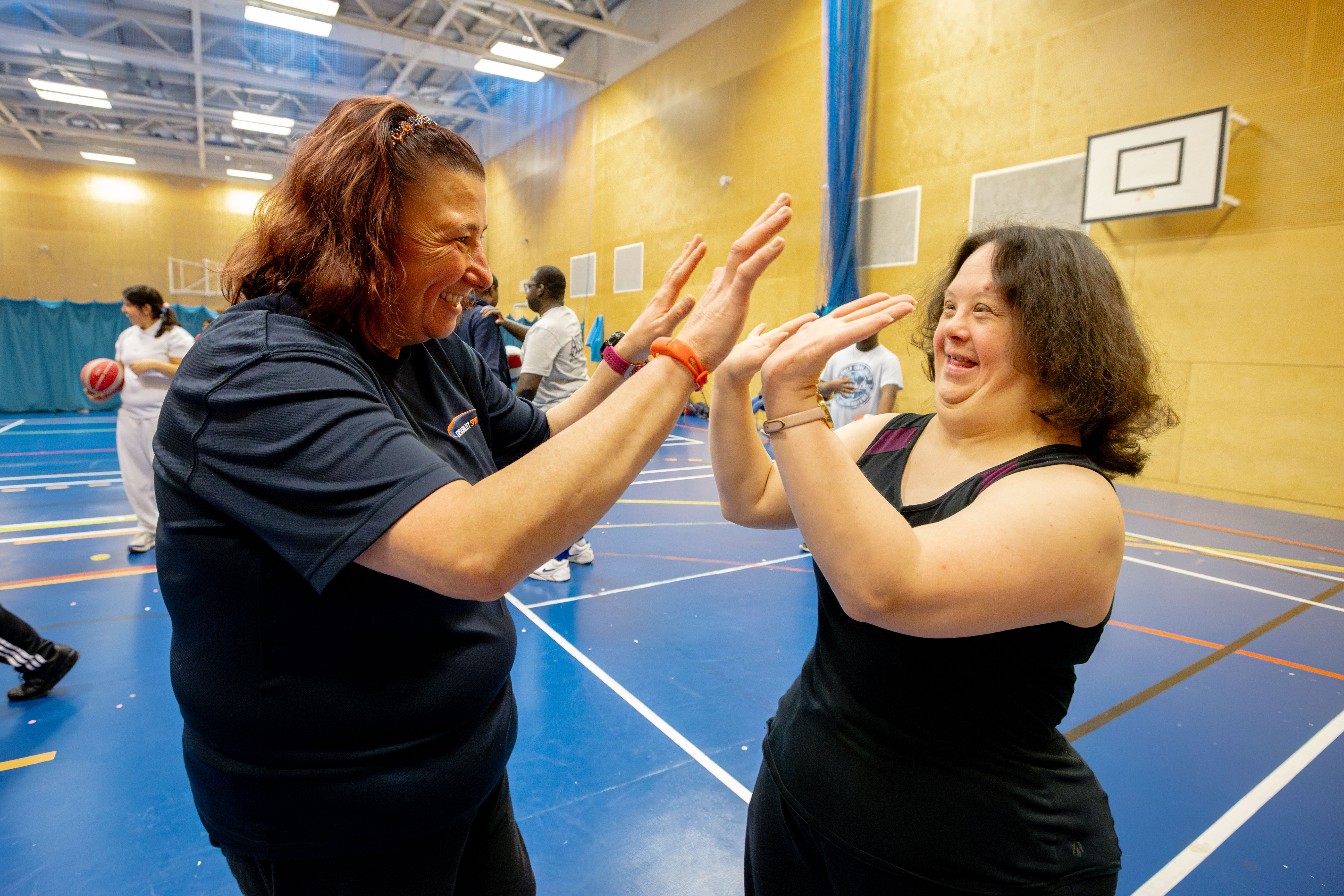 Two women laugh while high-fiving. They are in a sports hall with others playing sport behind them.